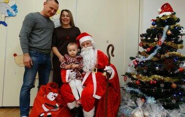 Sala zabaw - na pierwszym planie Mikołaj z laską, dzwoneczkiem w ręce i dziewczynką na kolanach siedzący na krześle. Tuż za nim stoją rodzice dziecka. W tle zielona choinka udekorowana błyszczącymi lampkami.