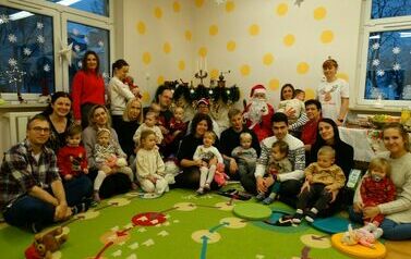 Sala zabaw - na pierwszym planie Mikołaj z laską i dzwoneczkiem w ręce siedzący na krześle. Obok niego dyrektor, rodzice z dziećmi na rękach i opiekunowie grupy. W tle kominek udekorowany zielonym łańcuchem.