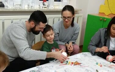 Sala zabaw - rodzice z dzieckiem siedzą przy stoliku, na kt&oacute;rym leżą kolorowe farby. Jedno z rodzic&oacute;w trzyma w ręce białą bombkę.W ręce trzymają