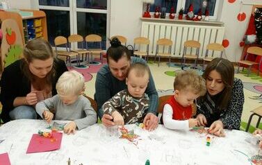 Sala zabaw - grupa rodzic&oacute;w z dziećmi siedzą przy stoliku. Dzieci trzymają w ręce pędzelki i dotykają nimi gwiazdek leżących na stole.