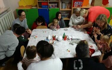 Sala zabaw - grupa rodzic&oacute;w z dziećmi siedzą przy stoliku, na kt&oacute;rym leżą białe bombki, kolorowe naklejki, pędzelki i farby do malowania.