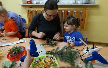 Sala zabaw - na pierwszym planie rodzic z dzieckiem siedzący przy stoliku, na kt&oacute;rym leżą zielone gałązki świerku, świeczki, szyszki i suszone cytrusy. 