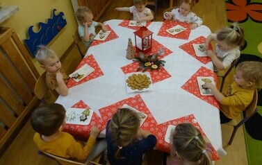 Sala zabaw-dzieci siedzą na krzesełkach przy stoliku, nakrytym białym obrusem i czerwonymi serwetkami. Na nich przed każdym dzieckiem stoją białe tacki z pierniczkami i kolorową posypką.
