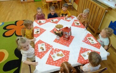 Sala zabaw-dzieci siedzą na krzesełkach przy stoliku, nakrytym białym obrusem i czerwonymi serwetkami. Na nich przed każdym dzieckiem stoją białe tacki z pierniczkami i kolorową posypką.