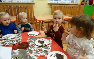 Sala zabaw-dzieci siedzą na krzesełkach przy stoliku, na kt&oacute;rym leżą talerzyki z brązowymi pierniczkami. Jedne dotykają rączkami pierniczk&oacute;w, inne paluszkami dotykają własnych ust.