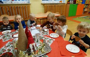 Sala zabaw-dzieci siedzą na krzesełkach przy stoliku, na kt&oacute;rym leżą talerzyki z brązowymi pierniczkami. Dzieci dotykają rączkami pierniczk&oacute;w i przykładają do ust.