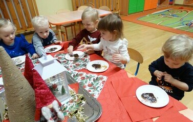 Sala zabaw-dzieci siedzą na krzesełkach przy stoliku, na kt&oacute;rym leżą talerzyki z brązowymi pierniczkami. Dzieci dotykają rączkami pierniczk&oacute;w. 