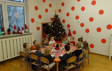 Sala zabaw-dzieci siedzą na krzesełkach przy stoliku, na kt&oacute;rym leżą pierniczki i małe torebki. Przed każdym dzieckiem stoi talerzyk z pierniczkiem i kolorową posypką. W tle błyszcząca duża choinka i dwa krasnale.