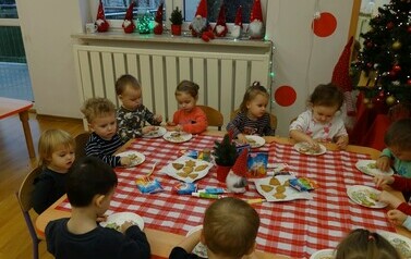 Sala zabaw - grupa dzieci siedzi na krzesełkach przy stoliku, na kt&oacute;rym leżą pierniczki, małe torebki i tubki. Przed każdym dzieckiem stoi talerzyk z pierniczkiem i kolorową posypką. Dzieci dotykają rączkami pierniczk&oacute;w.