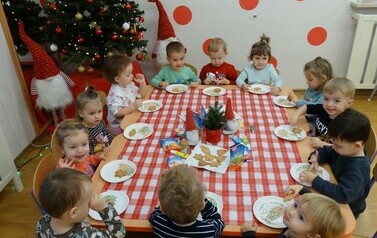 Sala zabaw-dzieci siedzą na krzesełkach przy stoliku, na kt&oacute;rym leżą pierniczki i kolorowe torebki. Przed każdym dzieckiem stoi talerzyk z pierniczkiem i kolorową posypką. W tle błyszcząca duża choinka i dwa krasnale.