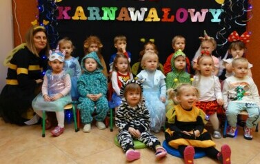 Na tle czarnej ścianki z kolorowym napisem "Bal Karnawałowy" grupa dzieci z opiekunem w strojach postaci z bajek. 