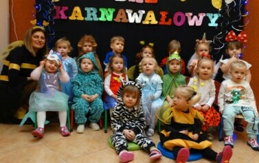 Na tle czarnej ścianki z kolorowym napisem "Bal Karnawałowy" grupa dzieci z opiekunem w strojach postaci z bajek. 