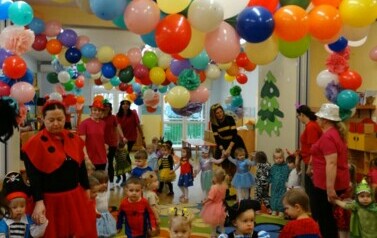 Sala zabaw-opiekunowie z grupą dzieci w strojach postaci z bajek trzymają się za ręce tworząc duże koło. W tle kolorowe balony.