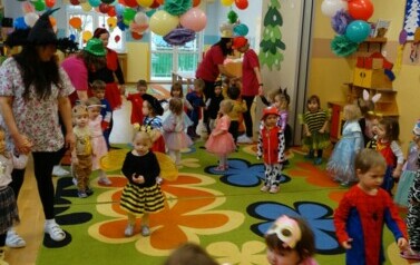 Sala zabaw-opiekunowie z grupą dzieci w strojach postaci z bajek trzymają się za ręce tworząc duże koło. W tle kolorowe balony.