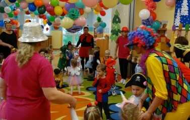Sala zabaw-opiekunowie z grupą dzieci w strojach postaci z bajek trzymają się za ręce tworząc duże koło. W tle kolorowe balony.
