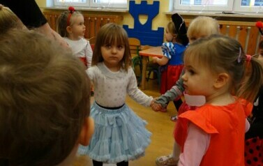 Sala zabaw-na pierwszym planie grupka dzieci w strojach postaci z bajek trzymająca się za ręce. W tle kolorowe balony.