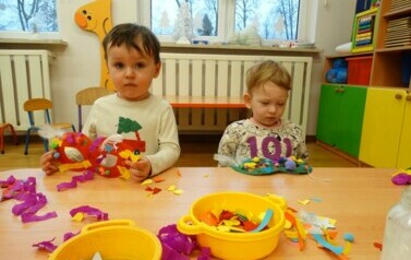 Sala zabaw-dw&oacute;jka dzieci siedzi przy stoliku, jedno z nich trzyma w ręce maskę karnawałową.