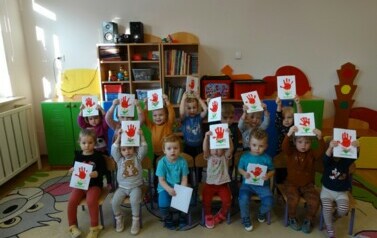 Sala zabaw - grupa dzieci siedzi na krzesełkach, w ręce trzyma laurki dla babci i dziadka.