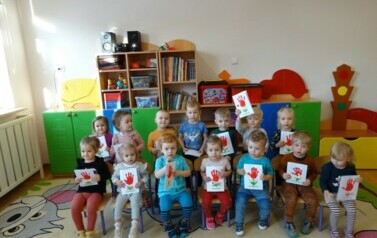 Sala zabaw - grupa dzieci siedzi na krzesełkach, w ręce trzyma laurki dla babci i dziadka.