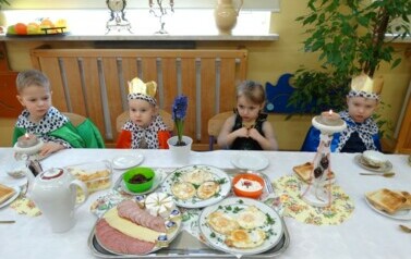 Sala zabaw-na zdjęciu długi st&oacute;ł, na nim biały obrus, zastawa porcelanowa i talerze z jedzeniem. Wok&oacute;ł niego siedzi grupa dzieci ubrana w stroje kr&oacute;lewskie.
