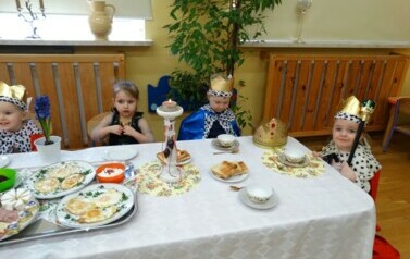 Sala zabaw-na zdjęciu st&oacute;ł, na nim biały obrus, zastawa porcelanowa i talerze z jedzeniem. Wok&oacute;ł niego siedzi grupka dzieci ubrana w stroje kr&oacute;lewskie.