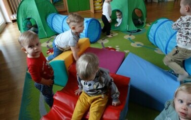 Sala zabaw-na podłodze stoją namioty, tunele i kolorowe piankowe klocki a wśr&oacute;d nich grupka dzieci.