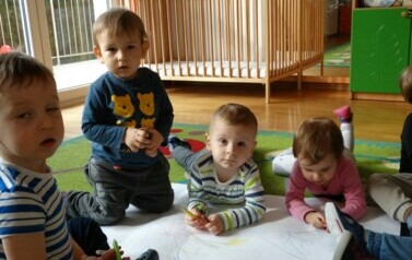 Sala zabaw- na podłodze leży biały duży karton a wok&oacute;ł niego grupka dzieci z kredkami w ręce.
