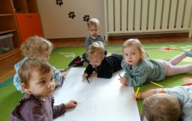 Sala zabaw- na podłodze leży biały duży karton a wok&oacute;ł niego grupka dzieci z kredkami w ręce.