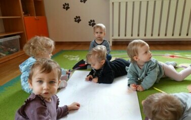 Sala zabaw- na podłodze leży biały duży karton a wok&oacute;ł niego grupka dzieci z kredkami w ręce.