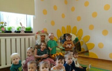 Sala zabaw-na dywanie siedzi grupka dzieci z Opiekunem w przebraniu Pani Wiosny. Obok niej stoi chłopiec z koszem ż&oacute;łtych kwiat&oacute;w w ręce.