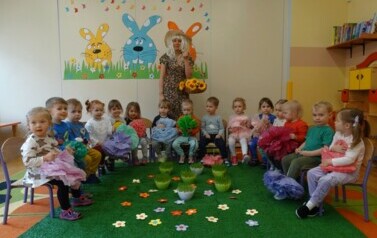 Sala zabaw-na tle dekoracyjnej ściany stoi Opiekun w przebraniu Pani Wiosny. W ręce trzyma kosz z ż&oacute;łtymi kwiatami. Przed nią siedzi grupa dzieci w kolorowych ubrankach.