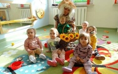 Sala zabaw-na dywanie siedzi grupka dzieci z opiekunem w przebraniu Pani Wiosny. Na kolanach trzyma dziecko a tuż przed nią stoi kosz z ż&oacute;łtymi kwiatami.