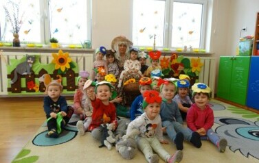 Sala zabaw-na dywanie siedzi grupka dzieci z opiekunem w przebraniu Pani Wiosny. Na kolanach trzyma dziecko z koszem ż&oacute;łtych kwiat&oacute;w w ręce. 