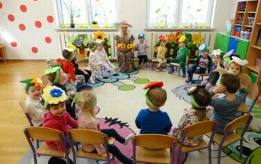 Sala zabaw-na krzesełku siedzi opiekun w przebraniu Pani Wiosny. Na kolanach trzyma kosz z ż&oacute;łtymi kwiatami. Wok&oacute;ł niej siedzi grupa dzieci z kolorowymi opaskami na czole.