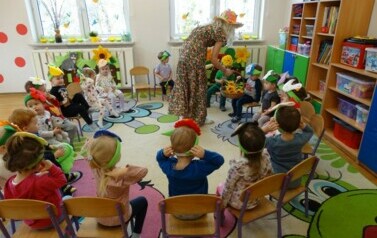 Sala zabaw-na krzesełkach siedzi grupa dzieci z kolorowymi opaskami na czole. Przy nich stoi schylony Opiekun w przebraniu Pani Wiosny z koszem ż&oacute;łtych kwiat&oacute;w.