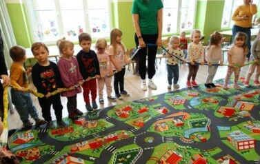 Sala zabaw-na dywanie stoi grupka dzieci z Opiekunem. W ręce trzymają kolorową gumę. 