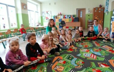 Sala zabaw-na dywanie siedzi grupka dzieci z Opiekunem. Na ich kolanach leży kolorowa guma. Dieci dotykają ją oburącz.