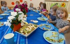 Grupka dzieci siedzi przy stoliku, w ręce trzymają łyżki i dotykają talerzy z warzywami.