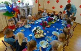 Grupa dzieci siedzi przy stoliku, na kt&oacute;rym stoją kolorowe kwiaty i salaterki z sałatką. W ręce trzymają łyżki, kt&oacute;rymi dotykają talerzy.