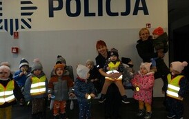 Grupa dzieci z opiekunem stoją na tle jasnej ściany, na kt&oacute;rej widnieje napis POLICJA. Opiekun i Pani policjantka trzymają dzieci na rękach.