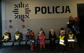 Grupa dzieci z opiekunem stoją na tle jasnej ściany, na kt&oacute;rej widnieje napis POLICJA.