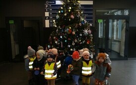 Grupa dzieci w odblaskowych ubrankach stoi na tle olbrzymiej zielonej choinki udekorowanej kolorowymi bombkami i odblaskami.