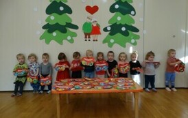 Sala zabaw - grupa dzieci trzyma w ręce laurki w kształcie serduszek. W tle jasna ściana a na niej babcia z dziadkiem i dwie choinki.