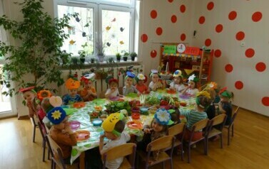 Sala zabaw-grupa dzieci siedzi przy stoliku, na kt&oacute;rym leżą świeże warzywa i owoce. Na ich głowach widnieją kolorowe opaski.