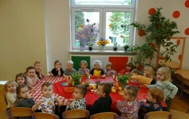 Sala zabaw-grupa dzieci siedzi przy stole nakrytym czerwonym obrusem. W tle z&oacute;łte kwiaty i drzewko z mandarynkami.
