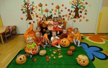 Sala zabaw - grupa uśmiechniętych dzieci w pomarańczowych ubrankach siedzi na dywanie, obok nich stoją dynie z wyciętymi otworami, w tle ściana z jesiennymi drzewkami, kolorowymi listkami i pomarańczowymi dyniami. 