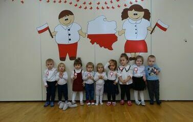 Sala zabaw - na tle jasnej ściany, udekorowanej symbolami narodowymi, stoi grupa dzieci w odświętnych ubraniach. Ich koszulki zdobią biało-czerwone kotyliony. 