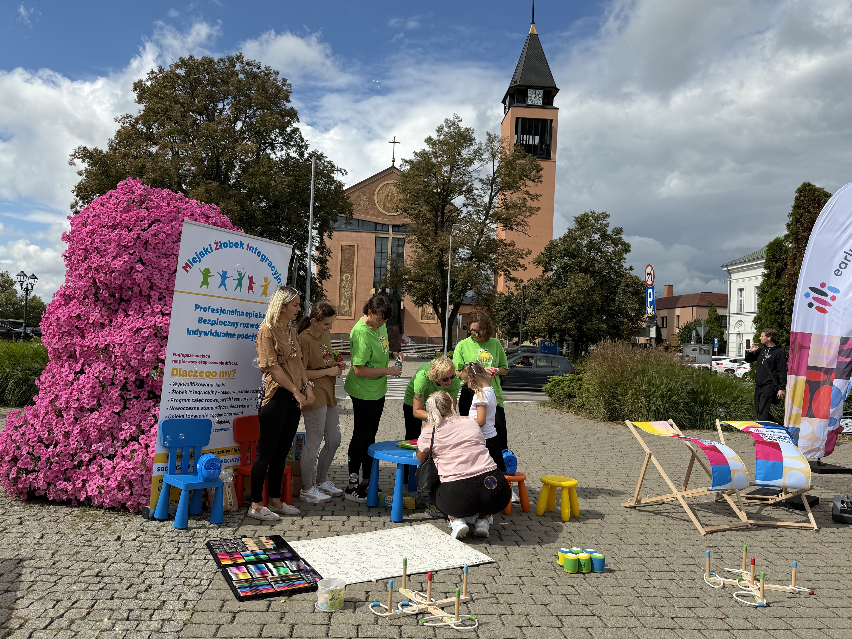 Na tle kolorowego plakatu stoją osoby dorosłe, tuż przed nimi kolorowy stolik z&nbsp;krzesełkami