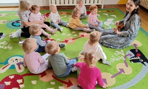 Zdjęcie przedstawia grupkę dzieci i osobę dorosłą siedzących na kolorowym dywanie.