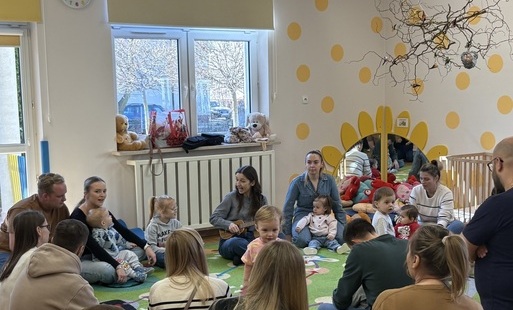 Grafika przedstawia grupę osób dorosłych wraz z dziećmi siedzących na dywanie w sali zabaw żłobka.